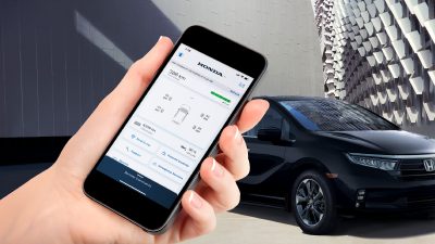 Closeup of hand holding a phone with HondaLink app open on screen. In the background, a black Honda Civic parked in front of modern architectural building.