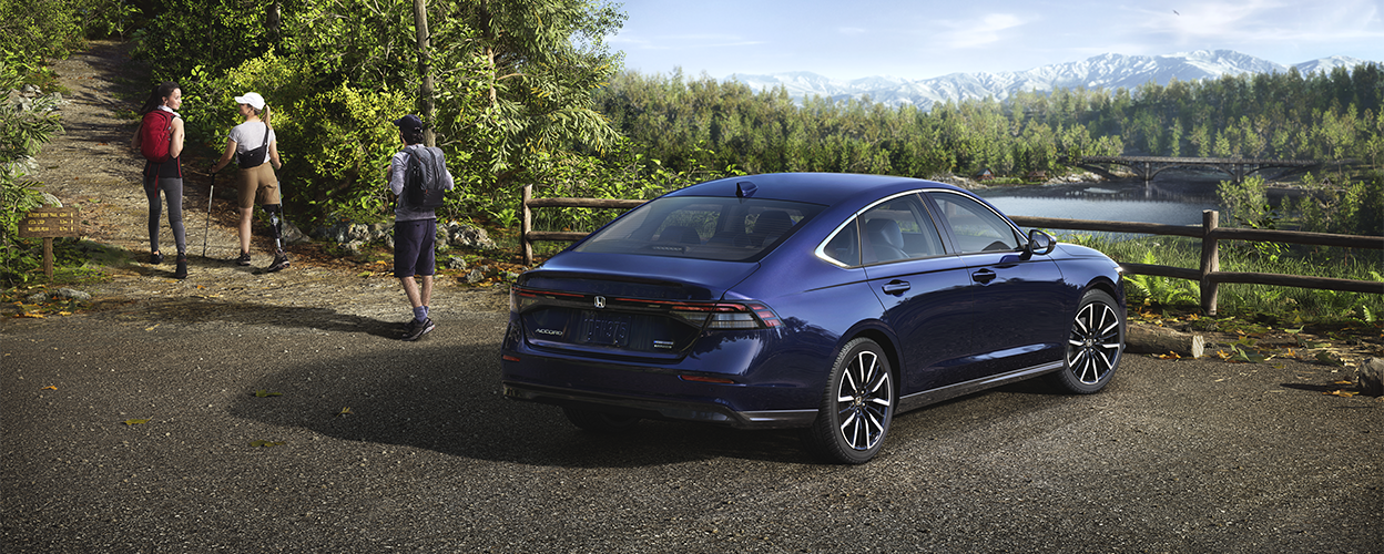 3/4 rear view of a parked blue Accord overseeing a forest and mountains. Three hikers walking away from it, embarking up a nearby hiking trail. 