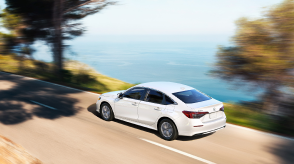 3/4 rear bird’s eye view of white Civic Sedan driving on an oceanside highway in the day.