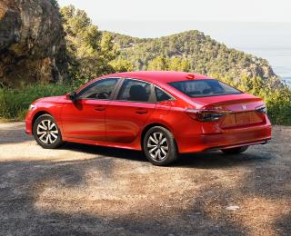 3/4 side rear view of red Civic Sedan parked at a scenic lookout spot overlooking plush green hills. A couple standing near it take in the scenery.