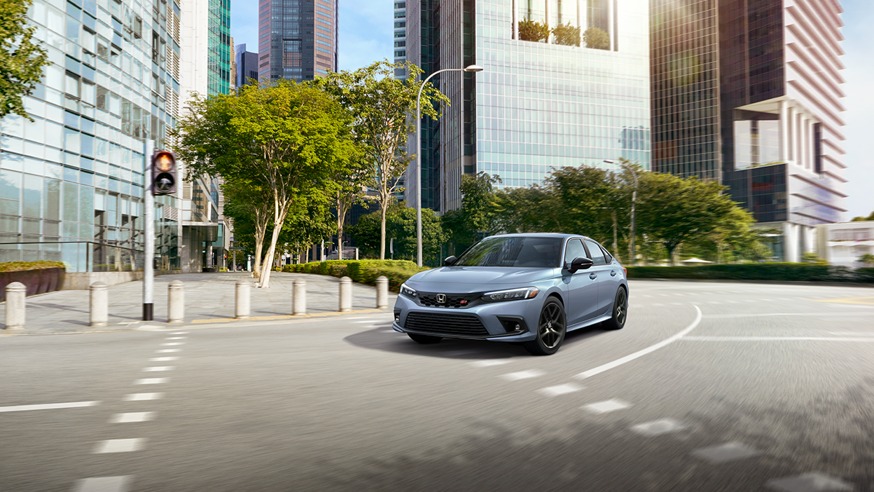 3/4 front view of greyish blue Civic Si taking a turn on a city street with lots of greenery on a sunny day.