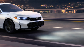 3/4 front side view of white Civic Si driving over a city bridge at night with headlights on. 