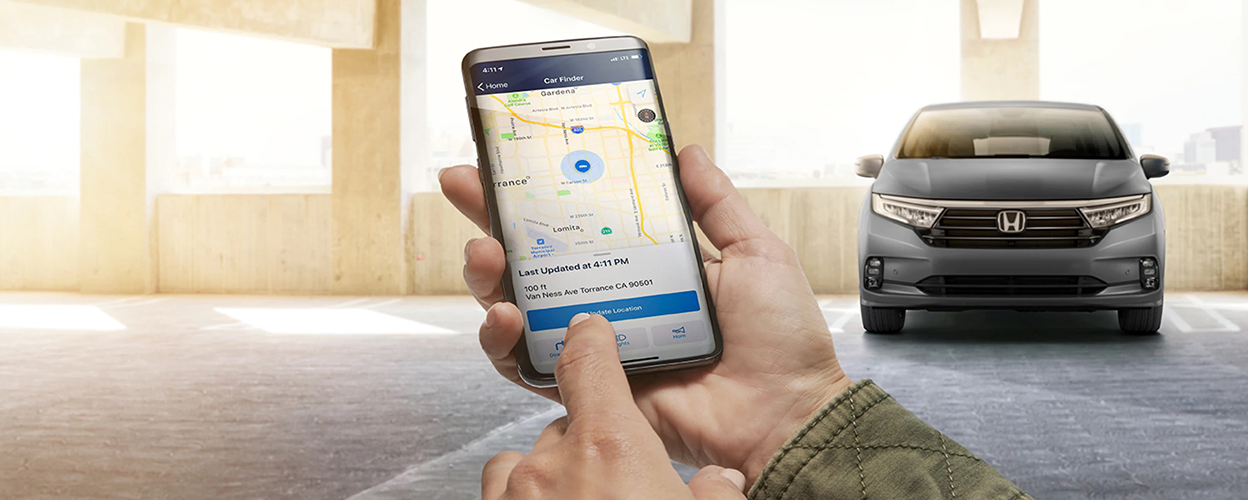 Hands using HondaLink app on phone. Grey odyssey parked in the background.