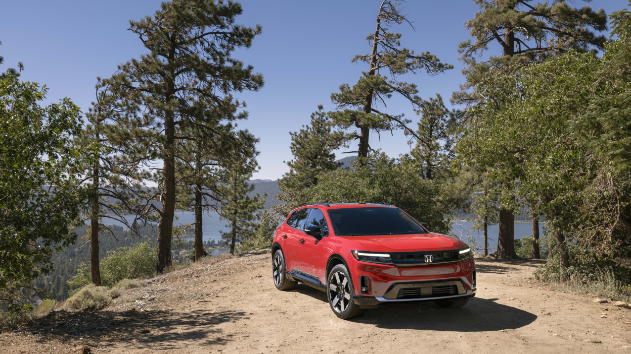 3/4 front view of red Prologue parked amongst trees in a forest. In the background we see a lake and mountians.