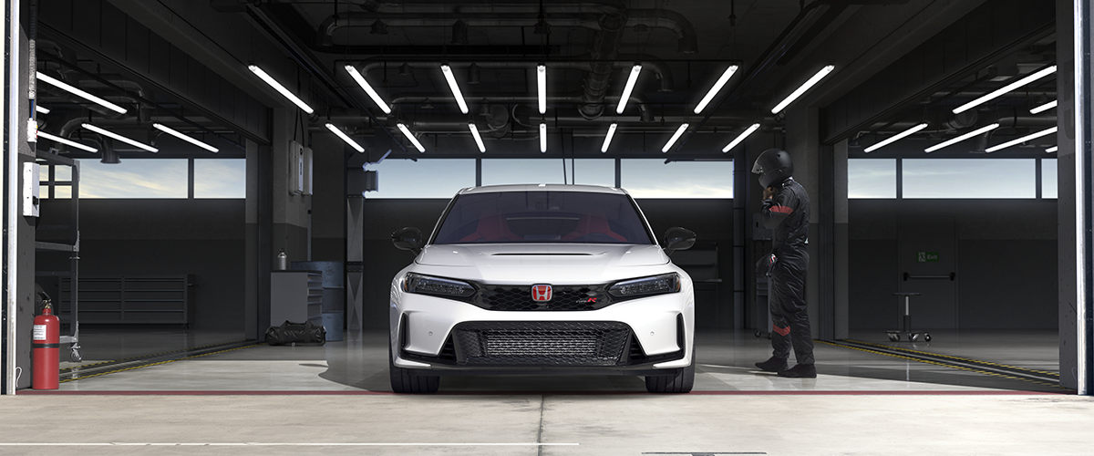 Front view of a white Type R parked in racing garage. A race car driver in black coveralls and helmet nears it. 